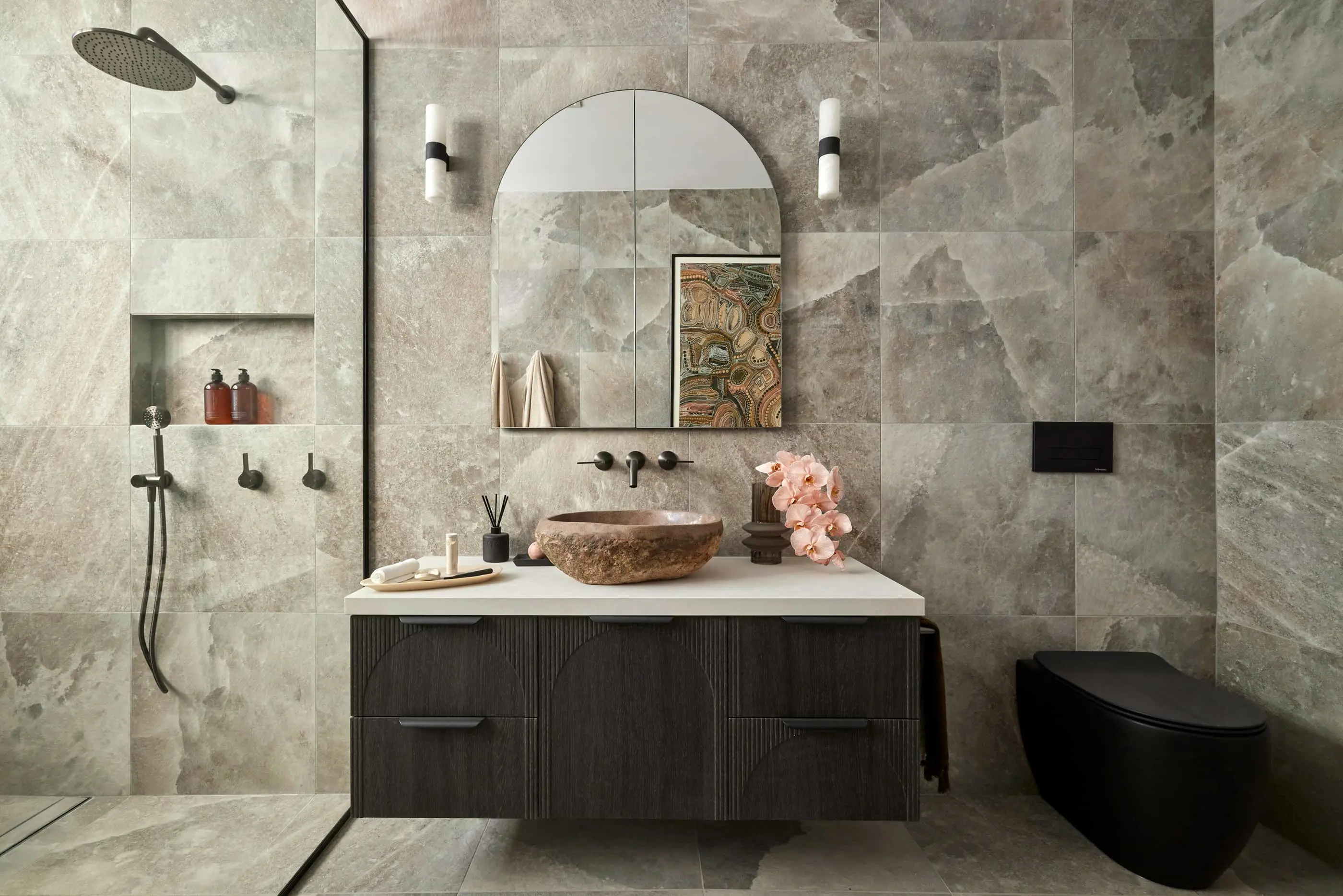Modern bathroom with stone walls, arched mirror, stone vessel sink on dark vanity, rainfall shower, and pink flowers.