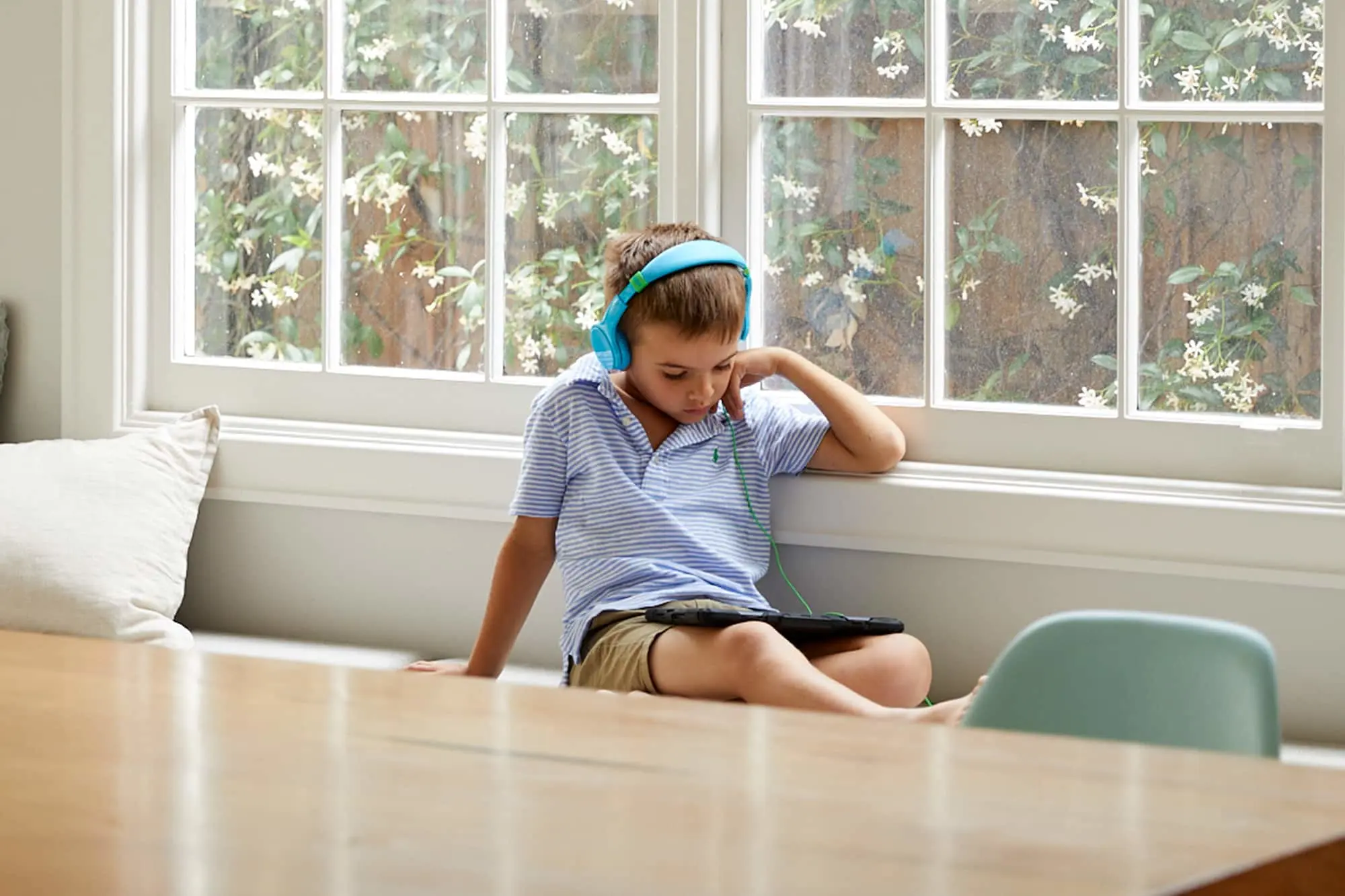 Child wearing blue headphones sitting by window with tablet, flowering plants visible outside.