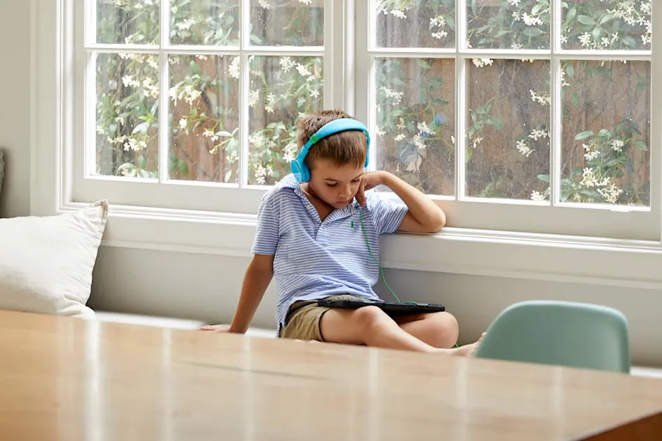 Child wearing blue headphones sitting by window with tablet, flowering plants visible outside.