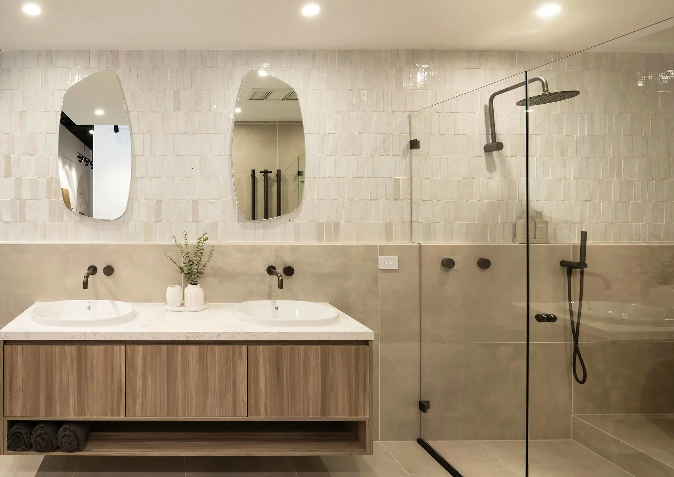 Modern bathroom with dual sinks, wooden vanity, cream tiled walls, oval mirrors, and glass shower enclosure with black fixtures.