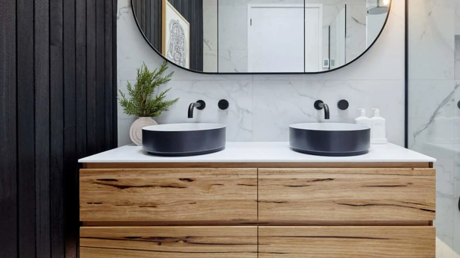 Modern bathroom with wooden vanity, two black vessel sinks, wall-mounted faucets, and oval mirror against marble wall.