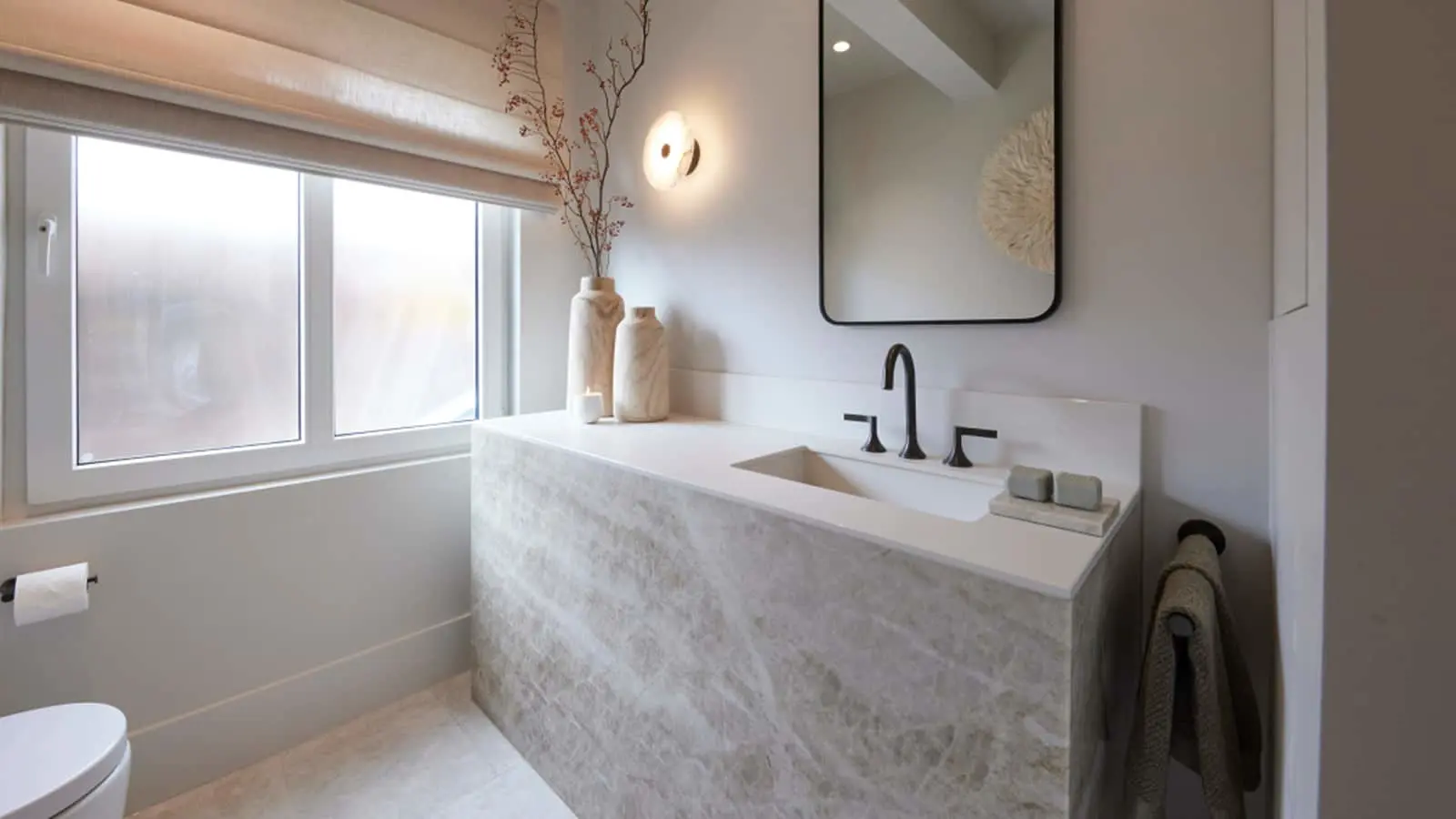 Modern minimalist bathroom with stone vanity, black fixtures, rectangular mirror, and decorative branches in a vase.