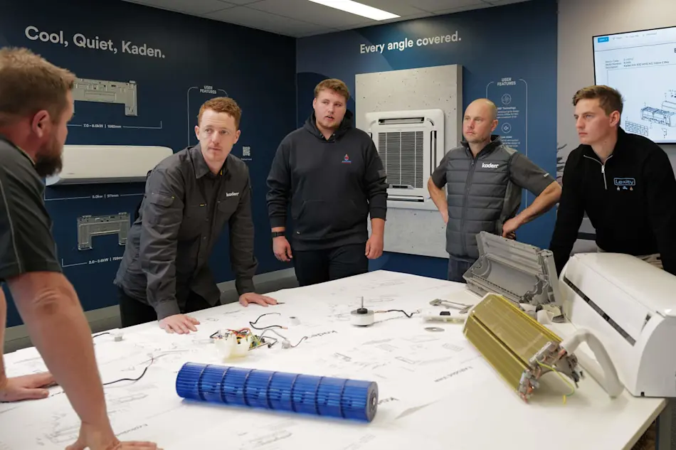 Group examining HVAC components on table in Kaden training room with wall-mounted air conditioning unit display.