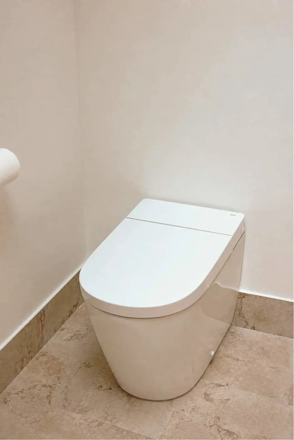 White modern smart toilet with closed lid in a bathroom corner with beige walls and stone tile flooring.