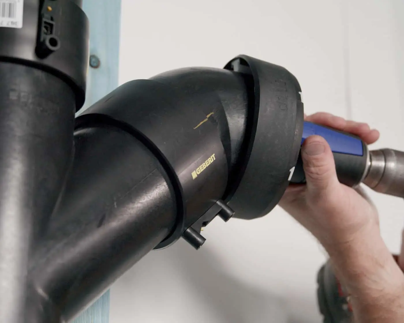 Hand using a power tool to connect black Geberit plumbing pipes with an elbow joint.