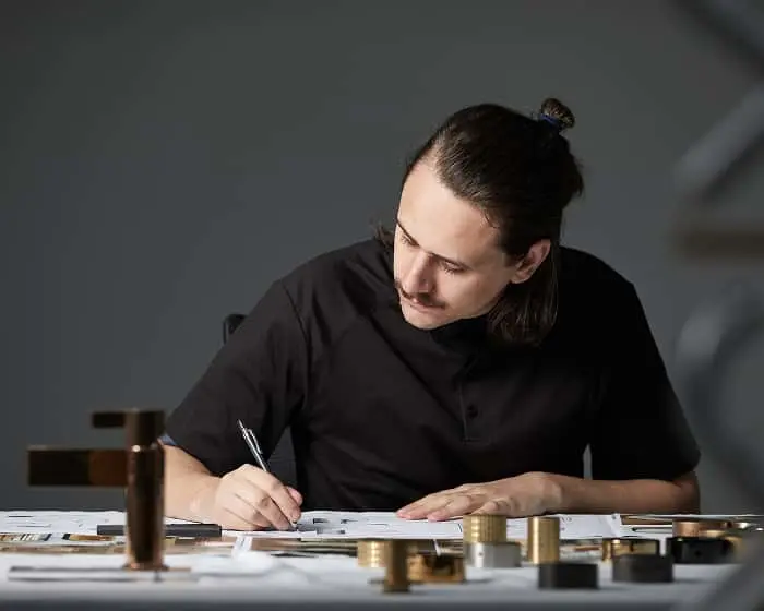 Person with hair in bun wearing black shirt writing in notebook at desk with brass objects and materials nearby.