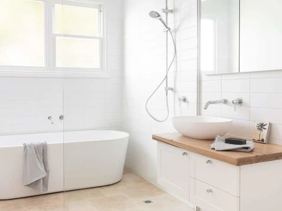 Modern white bathroom with freestanding tub, vessel sink on wooden countertop, glass shower, and natural light from window.