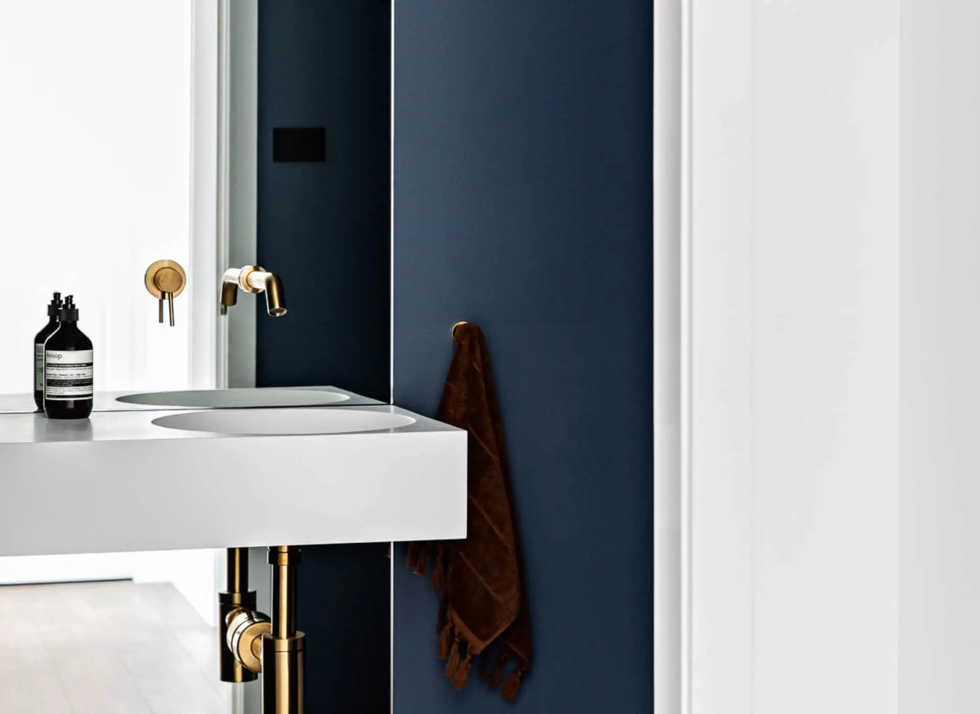 Modern bathroom with white sink, brass fixtures, navy blue wall, and brown towel hanging beside a mirror.