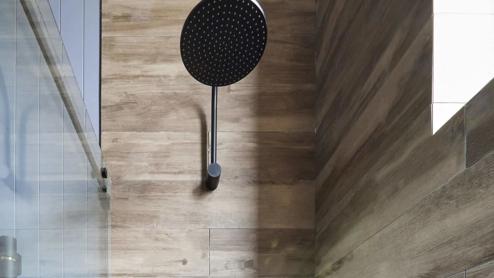 Modern bathroom with round black rainfall showerhead mounted on wood-look tile wall next to glass shower door.