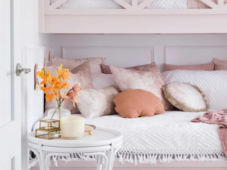 Pastel bedroom with pink pillows on white bed, orange flowers and candle on round white side table.