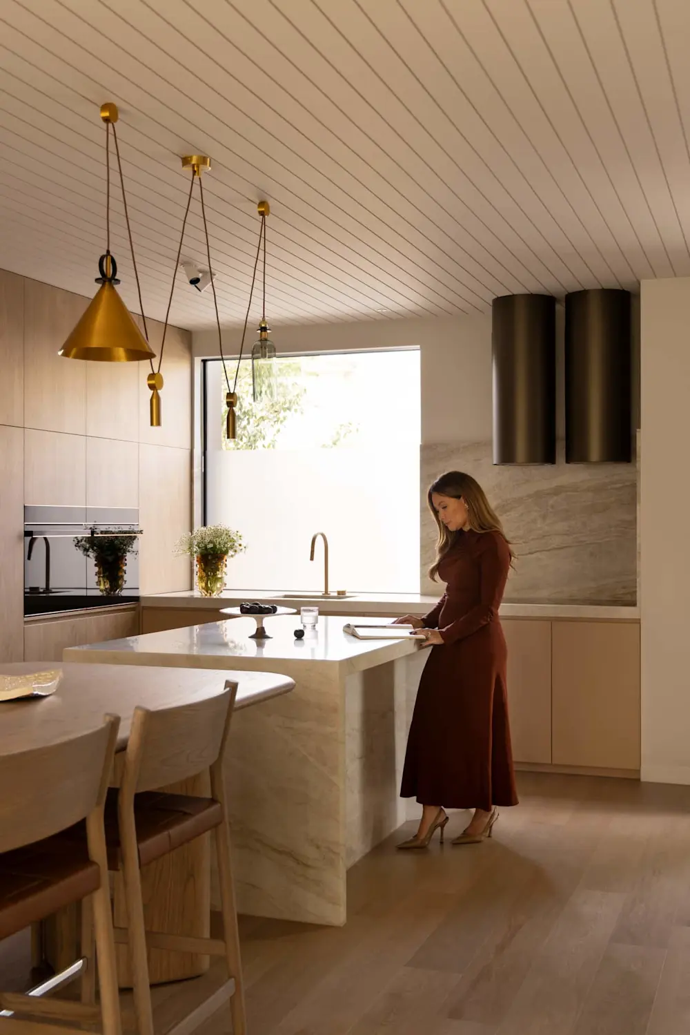 Woman in burgundy dress standing at marble kitchen island with gold pendant lights and wooden ceiling panels above.