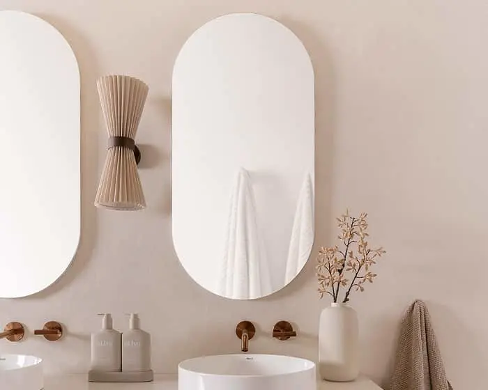 Minimalist bathroom with oval mirrors, white sink, beige wall sconce, and decorative vase with dried flowers.