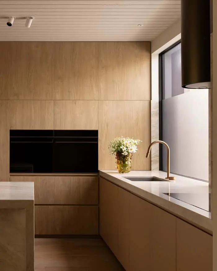 Modern kitchen with wooden cabinetry, white countertops, brass faucet, and small vase of daisies by a window.