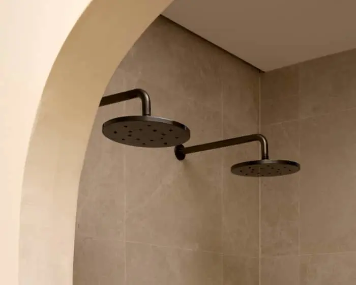Two gunmetal rainfall showerheads mounted on curved arms in a beige tiled shower alcove with an arched entrance.