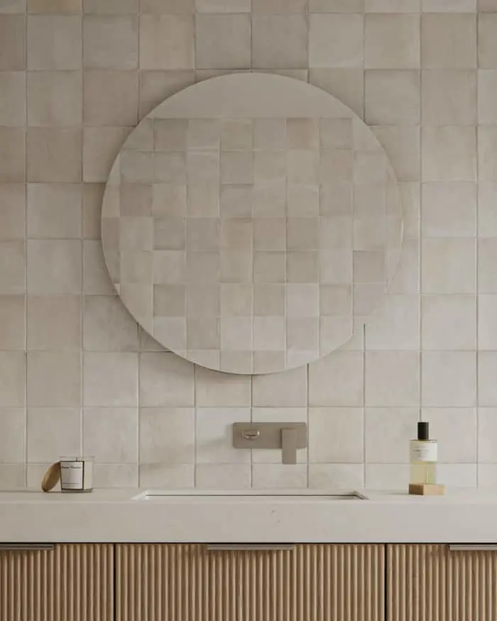 Round mirror above bathroom sink with beige tiled wall, wooden cabinet below, and minimal toiletries on counter.