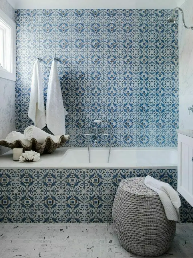 Bathroom with blue patterned tiles, white bathtub, hanging towels, decorative shell, and woven basket stool.
