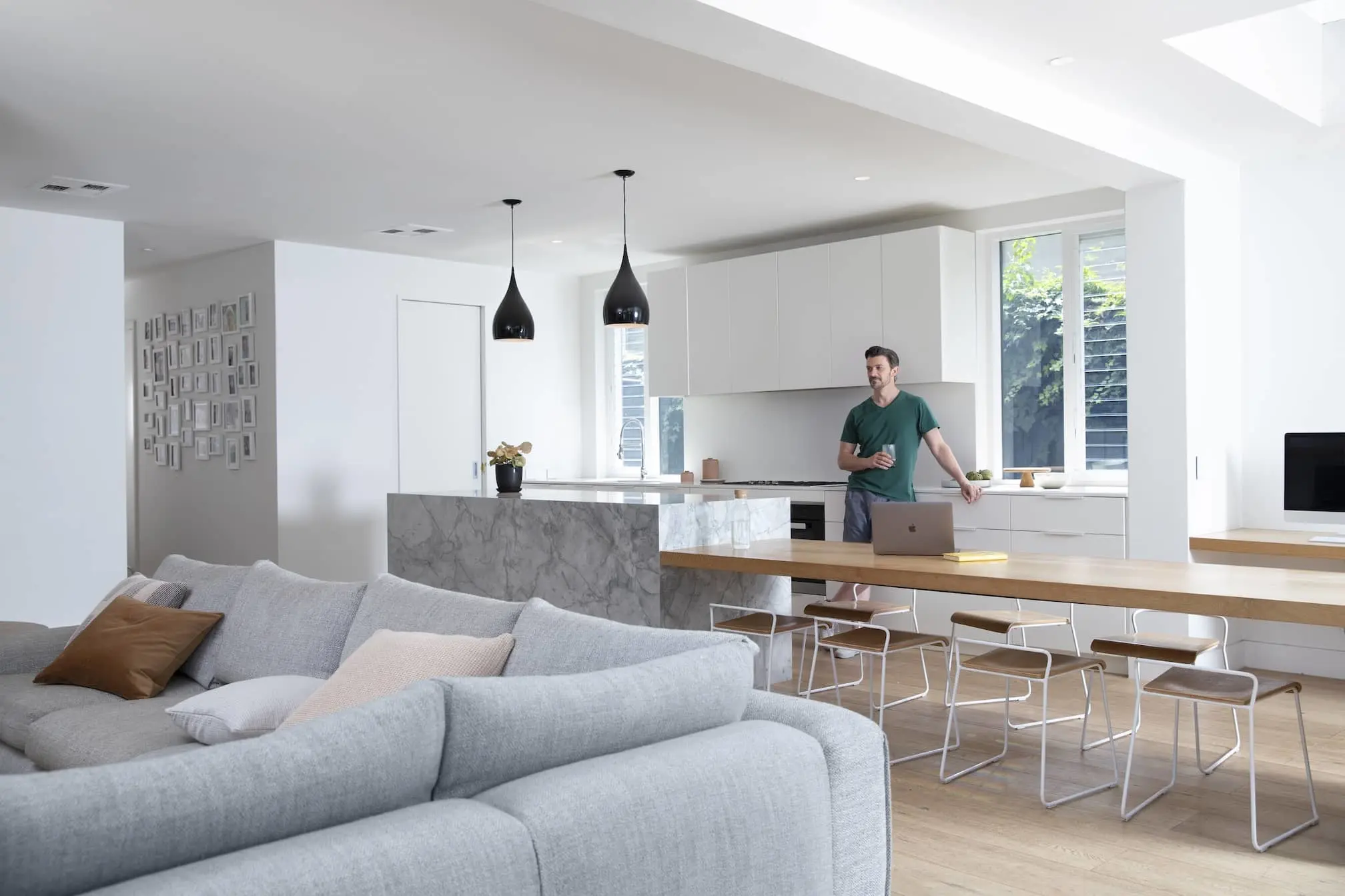 Modern open-concept kitchen and living room with gray sofa, marble island, and person in green shirt standing by laptop.