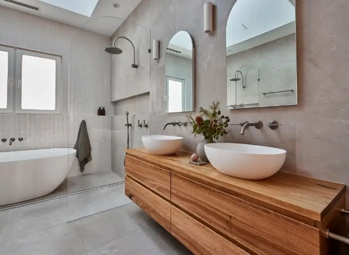 Modern bathroom with wooden vanity, white vessel sinks, arched mirrors, and freestanding bathtub in neutral tones.