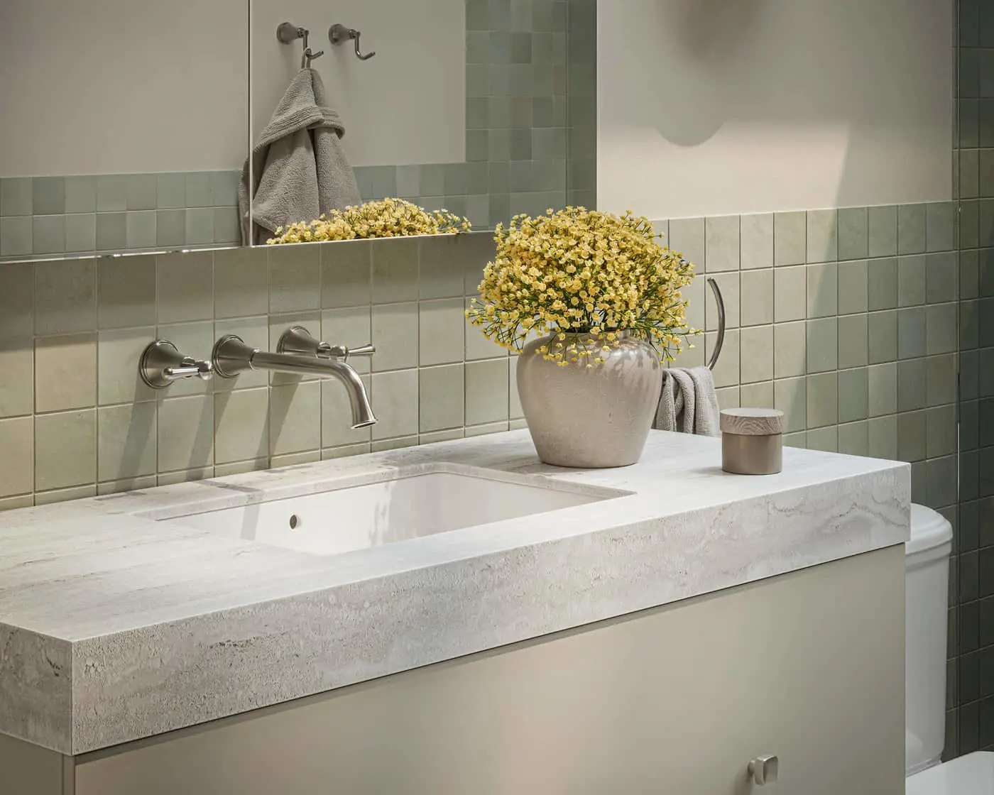 Bathroom vanity with white stone countertop, wall-mounted faucet, yellow flowers in vase, and green tile backsplash.