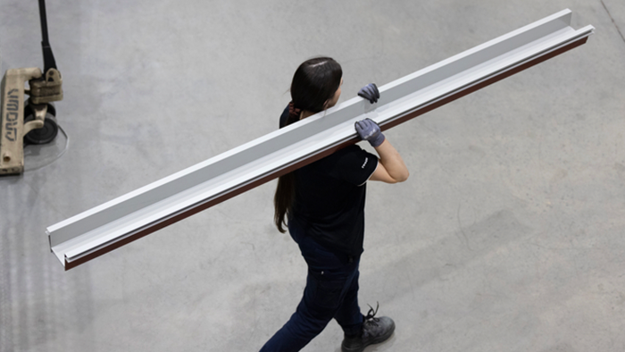 Person in dark clothing carrying a long metal beam or extrusion across a concrete floor in a warehouse.