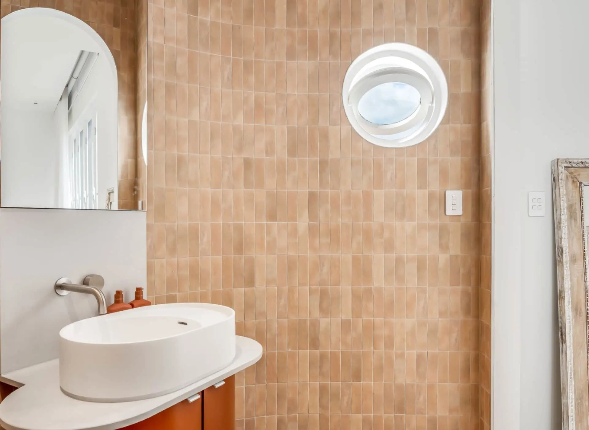 Modern bathroom with peach tiled wall, white vessel sink, circular window, and arched doorway.