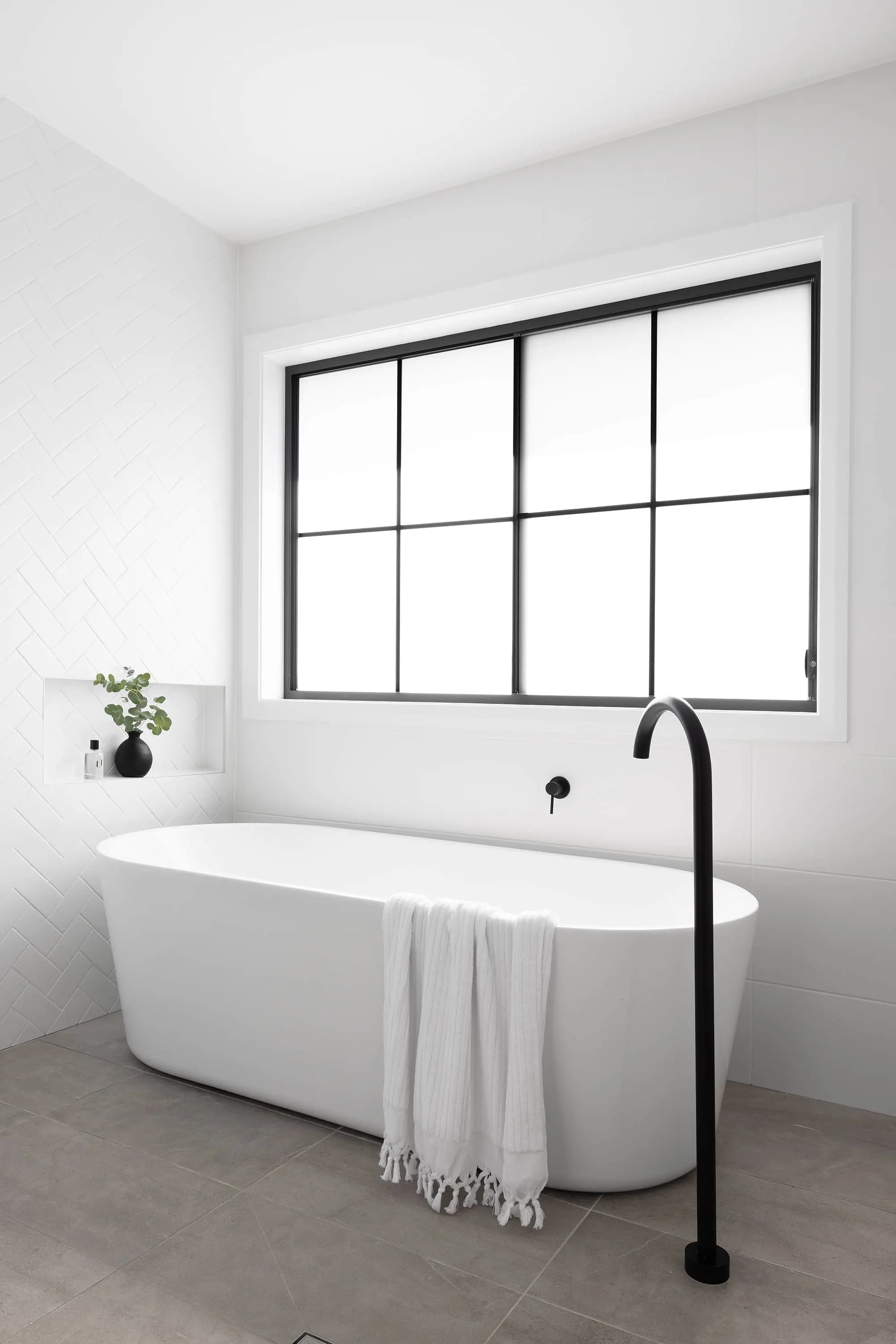 Modern white bathroom with freestanding tub, black faucet, window with black frame, and small plant in wall niche.