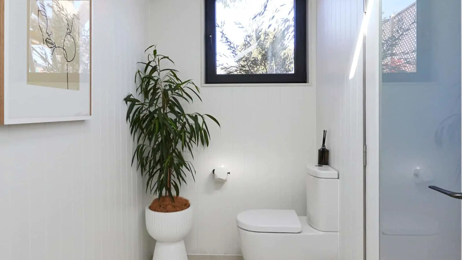 Modern white bathroom with toilet, potted palm plant, window with natural light, and minimalist decor.
