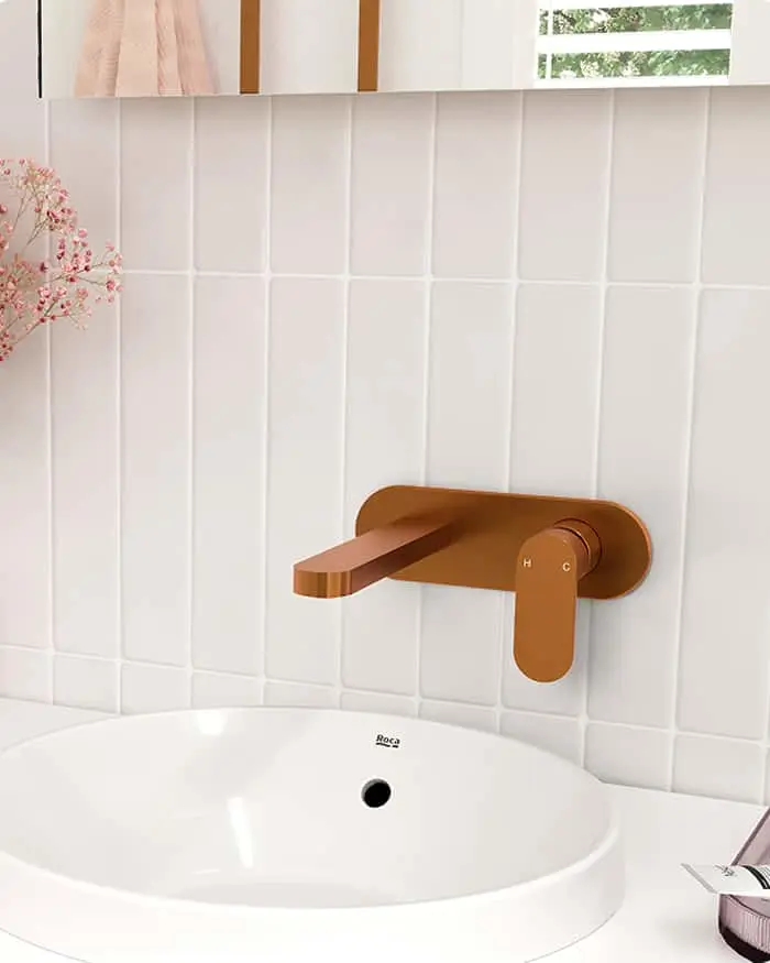 Bronze wall-mounted faucet against white vertical tiles, with white sink basin and pink floral decoration in bathroom.