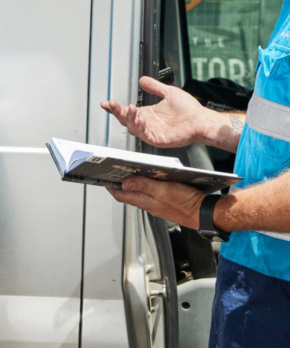Worker in blue safety vest holding a notebook while gesturing at a vehicle door.