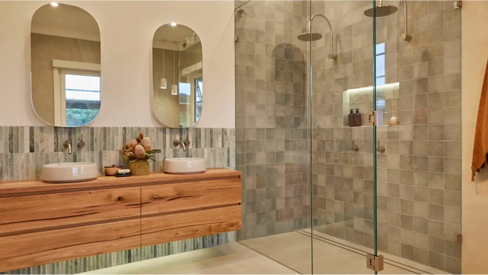 Modern bathroom with wooden vanity, dual vessel sinks, oval mirrors, and glass shower with gray tile walls.