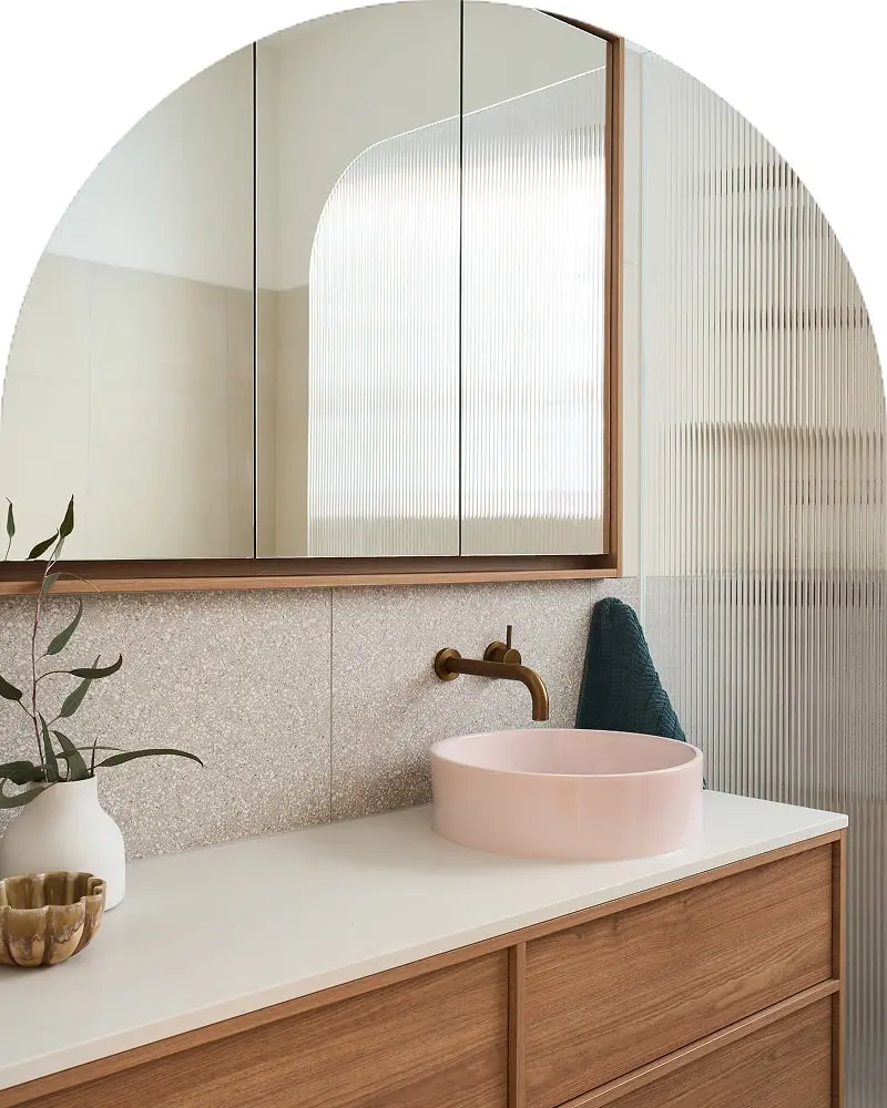 Modern bathroom with pink vessel sink, wooden vanity, arched mirror, brass faucet, and textured glass partition.