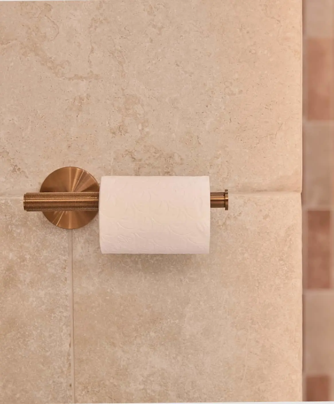 Copper toilet paper holder mounted on beige stone bathroom wall with white toilet paper roll.
