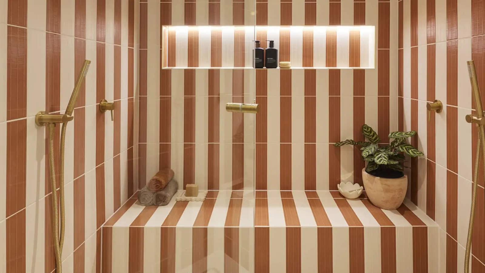 Modern shower with brown and cream striped tiles, brass fixtures, built-in shelf with toiletries, rolled towels, and a plant.