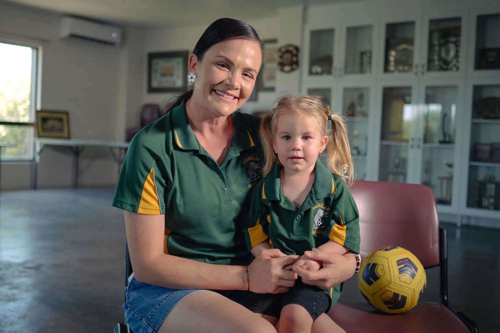 Adult and child wearing matching green and yellow sports uniforms sitting together with soccer ball nearby.