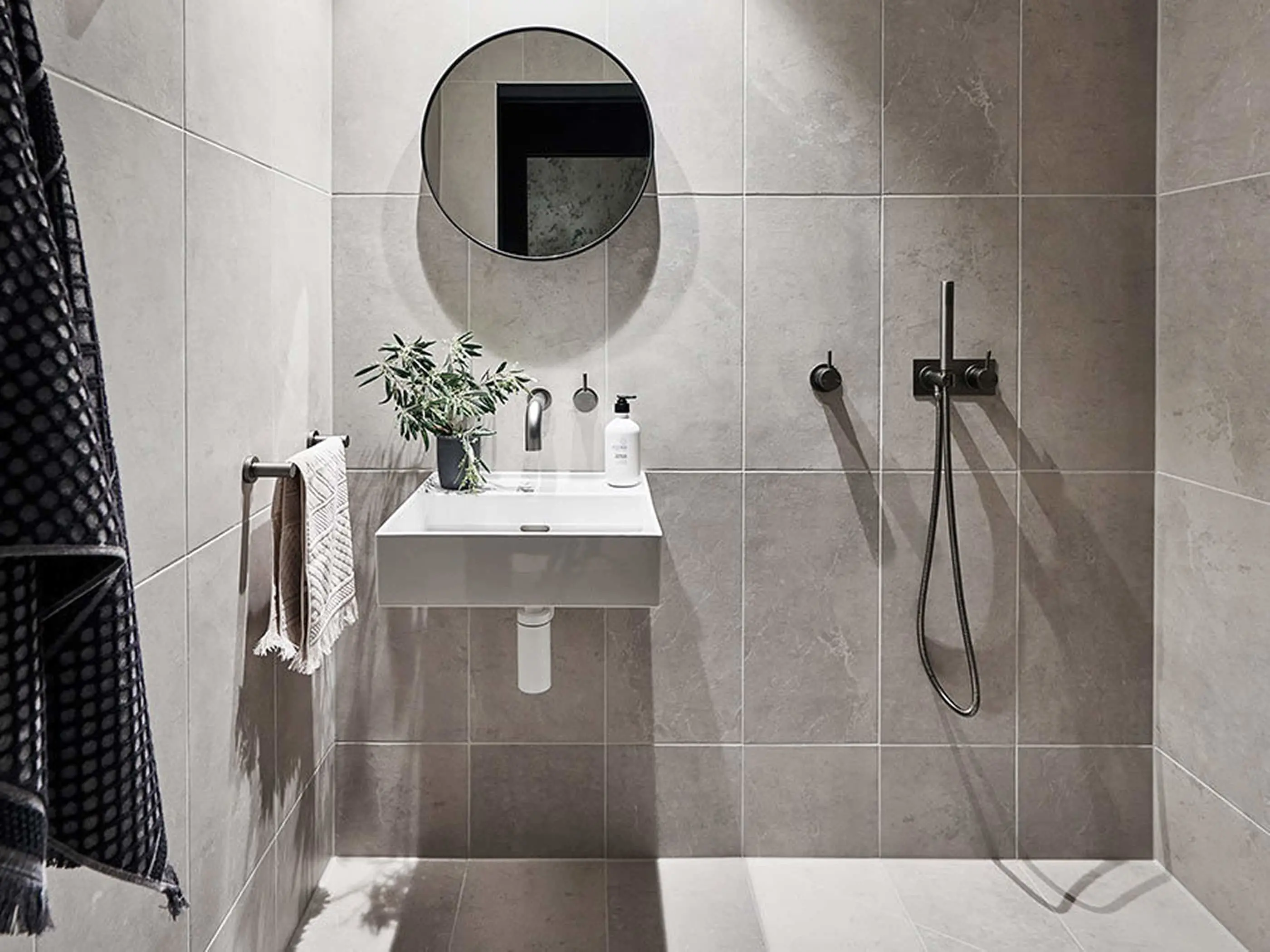Modern bathroom with gray tile walls, round mirror, white sink with small plant, and black shower fixtures.