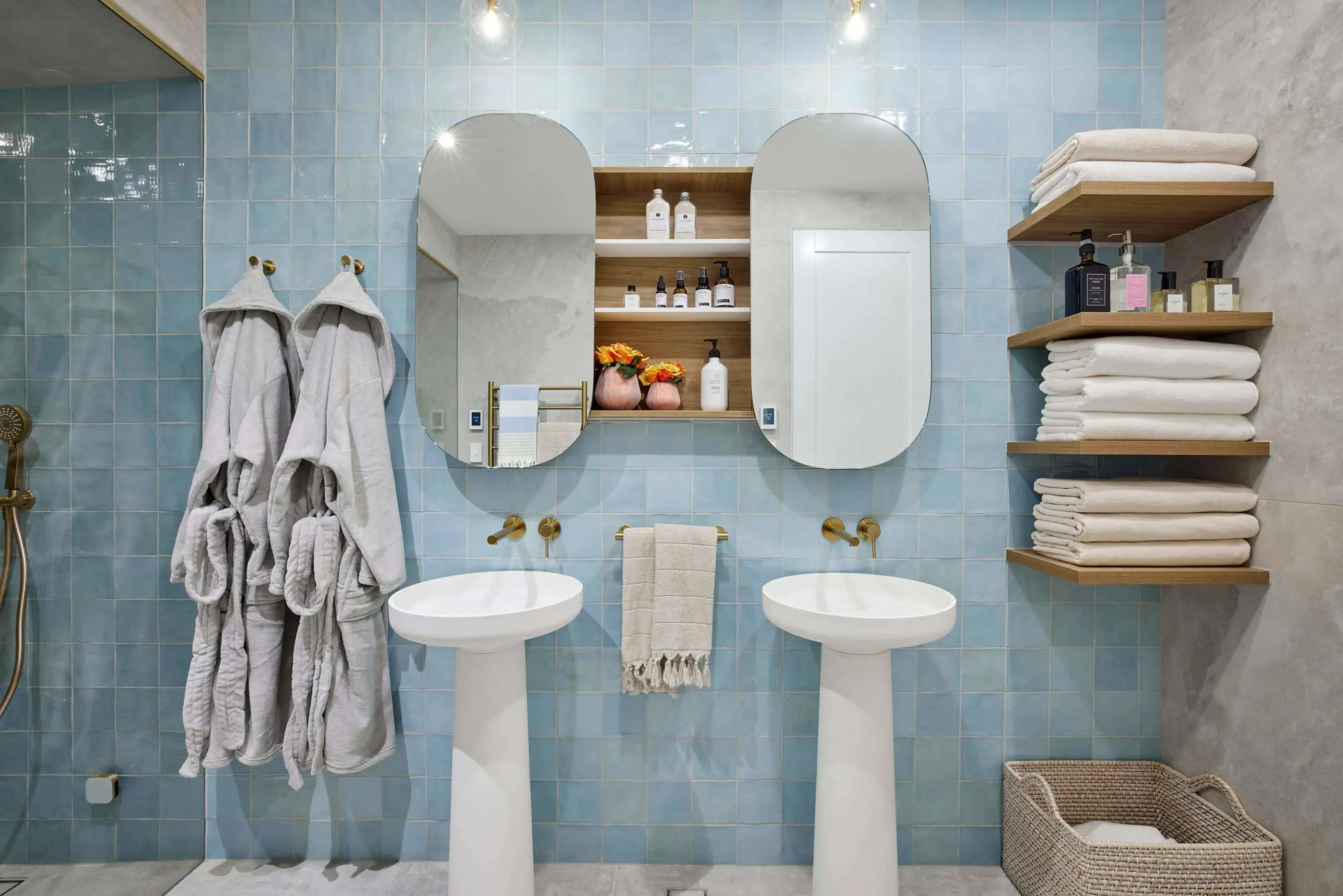 Modern bathroom with light blue tiles, dual pedestal sinks, mirrored cabinet, white robes, and wooden shelves with towels.