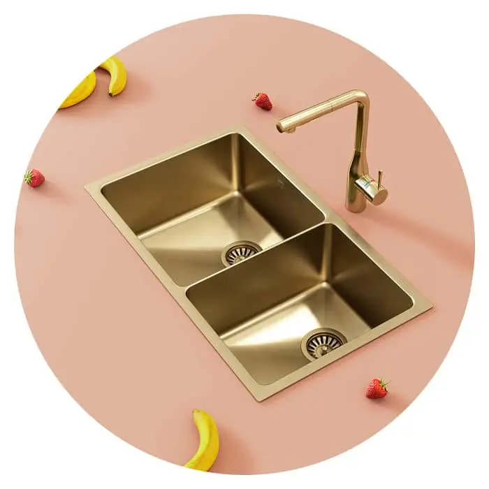 Gold double kitchen sink with matching faucet on pink surface, decorated with bananas and strawberries.