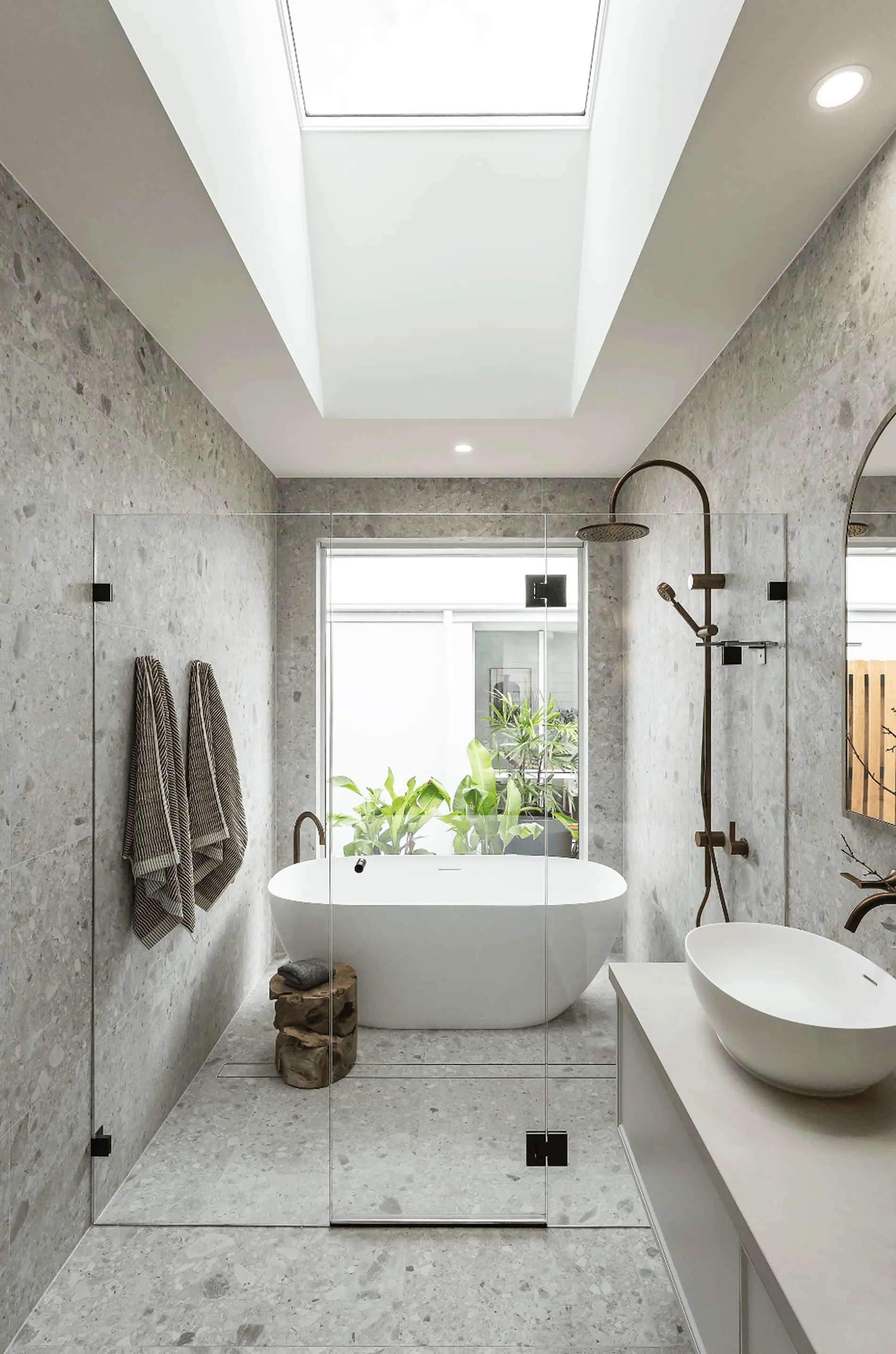 Modern bathroom with concrete walls, white freestanding tub, skylight, glass shower enclosure, and plants visible outside.