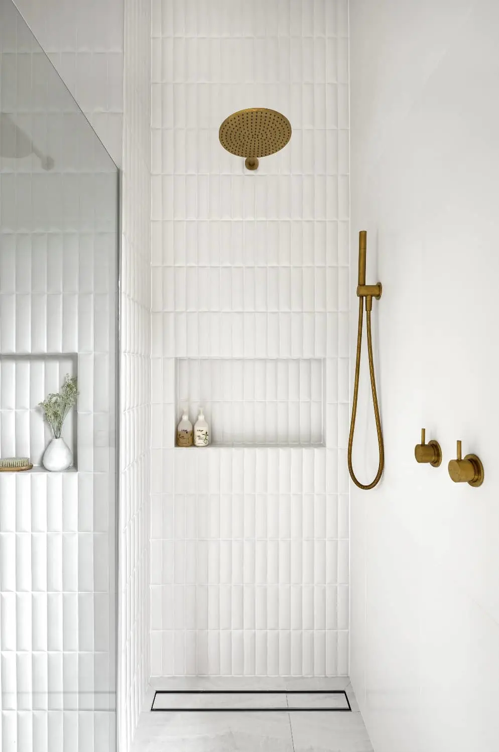 Modern white tiled shower with brass rainfall showerhead, handheld sprayer, and built-in niche for toiletries.