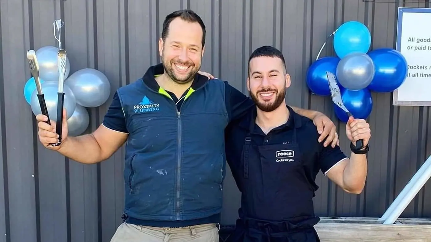Two smiling tradesmen in work uniforms holding BBQ utensils with blue and silver balloons decorating a gray wall behind them.