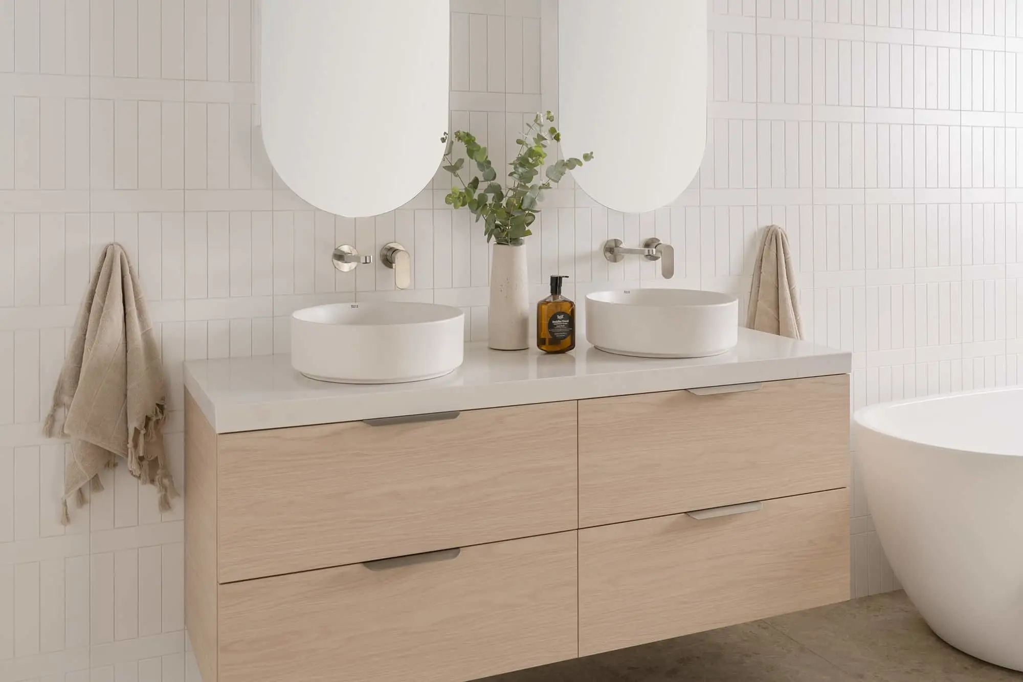 Modern bathroom with double white vessel sinks on light wood vanity, round mirrors, and eucalyptus in white vase.