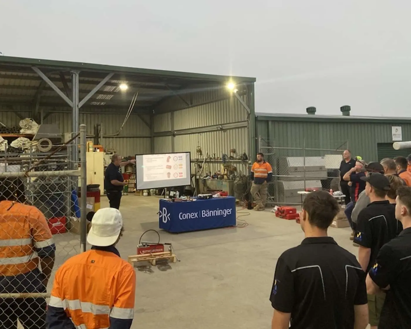 Workers in high-visibility gear attend a Conex Bänninger presentation in an industrial warehouse with projection screen.