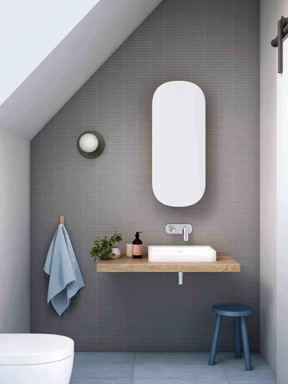 Modern bathroom with gray tile walls, wooden vanity, oval mirror, blue towel, small plant, and blue stool.