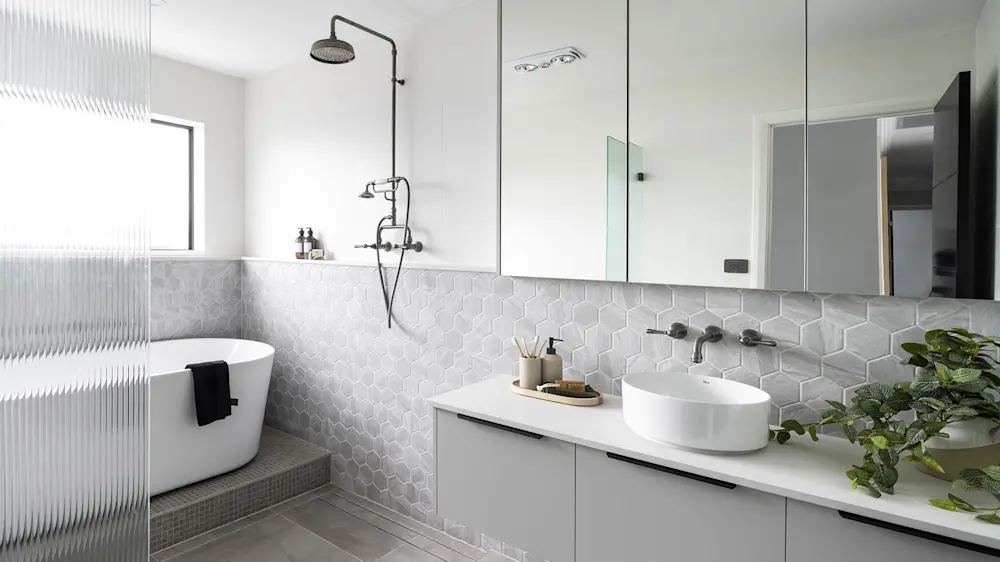 Modern white bathroom with hexagonal tile backsplash, freestanding tub, rainfall shower, vessel sink, and green plant.