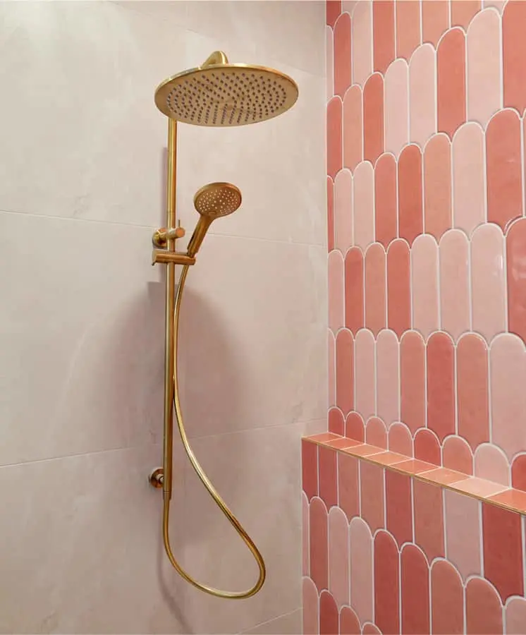 Gold rainfall showerhead with handheld attachment mounted on white wall next to pink scalloped tile in bathroom.