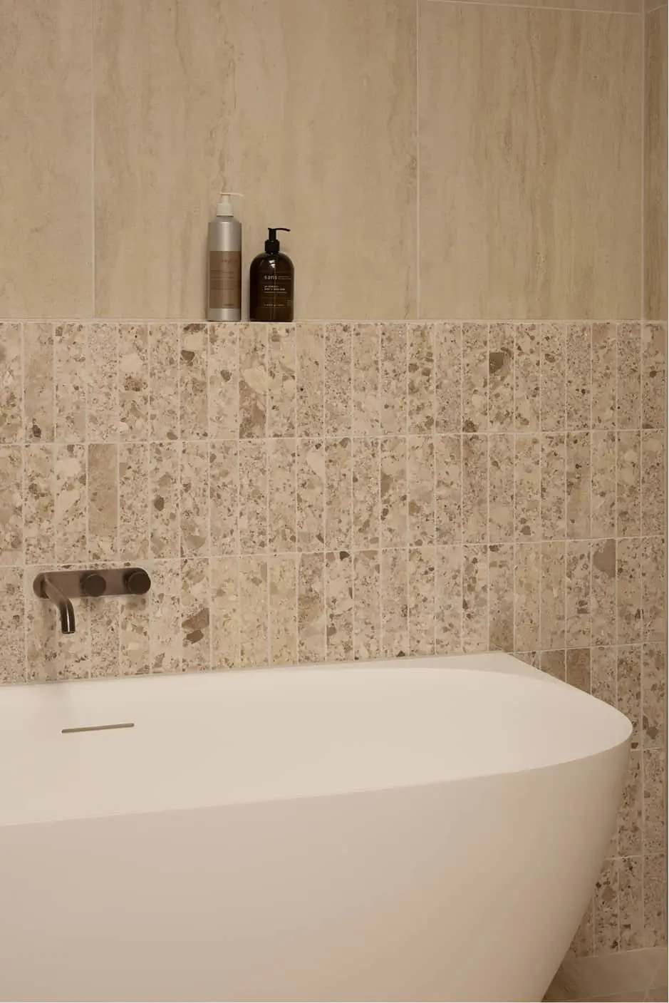 White bath with bronze wall-mounted faucet against beige terrazzo and marble tiled bathroom wall with soap bottles.