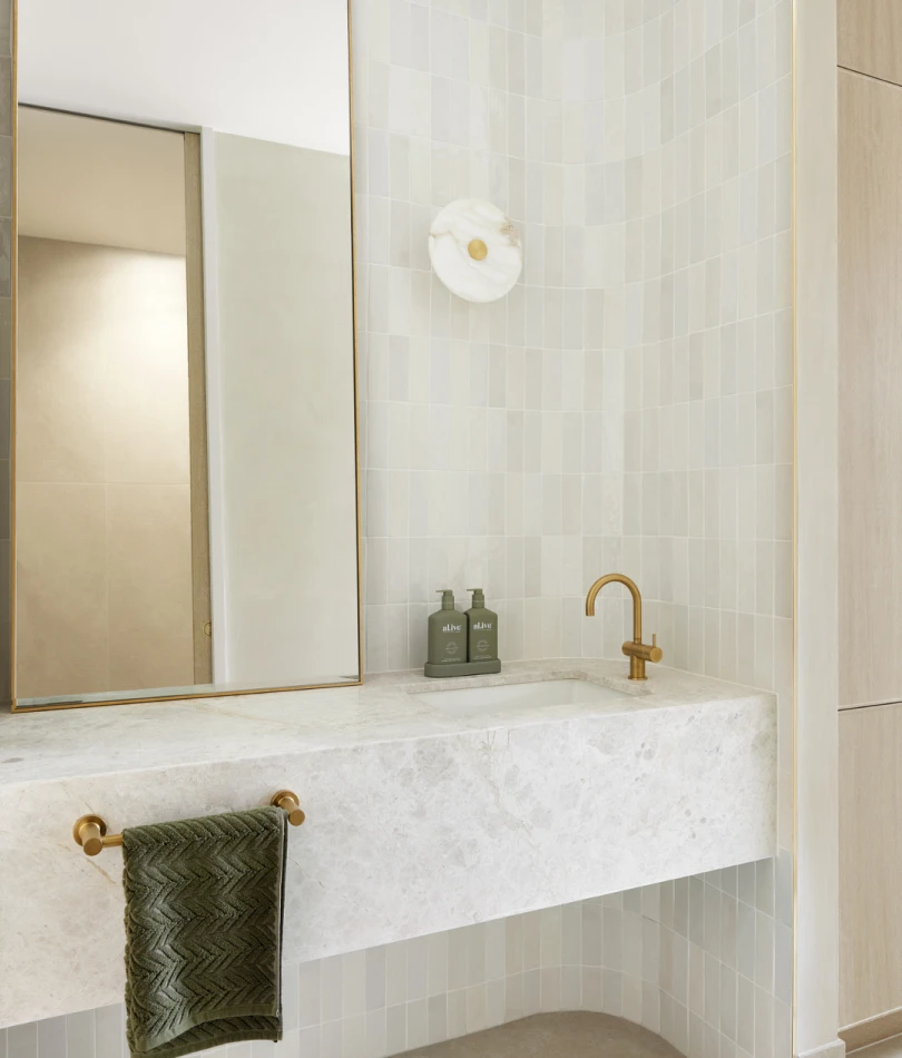 Modern bathroom with marble sink, gold fixtures, green towel, and light green tiled walls with circular wall sconce.