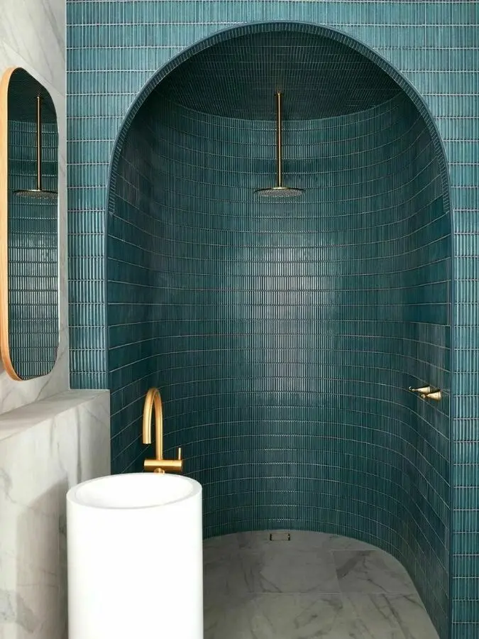 Modern bathroom with teal tile shower alcove, white pedestal sink, gold fixtures, and marble floor.