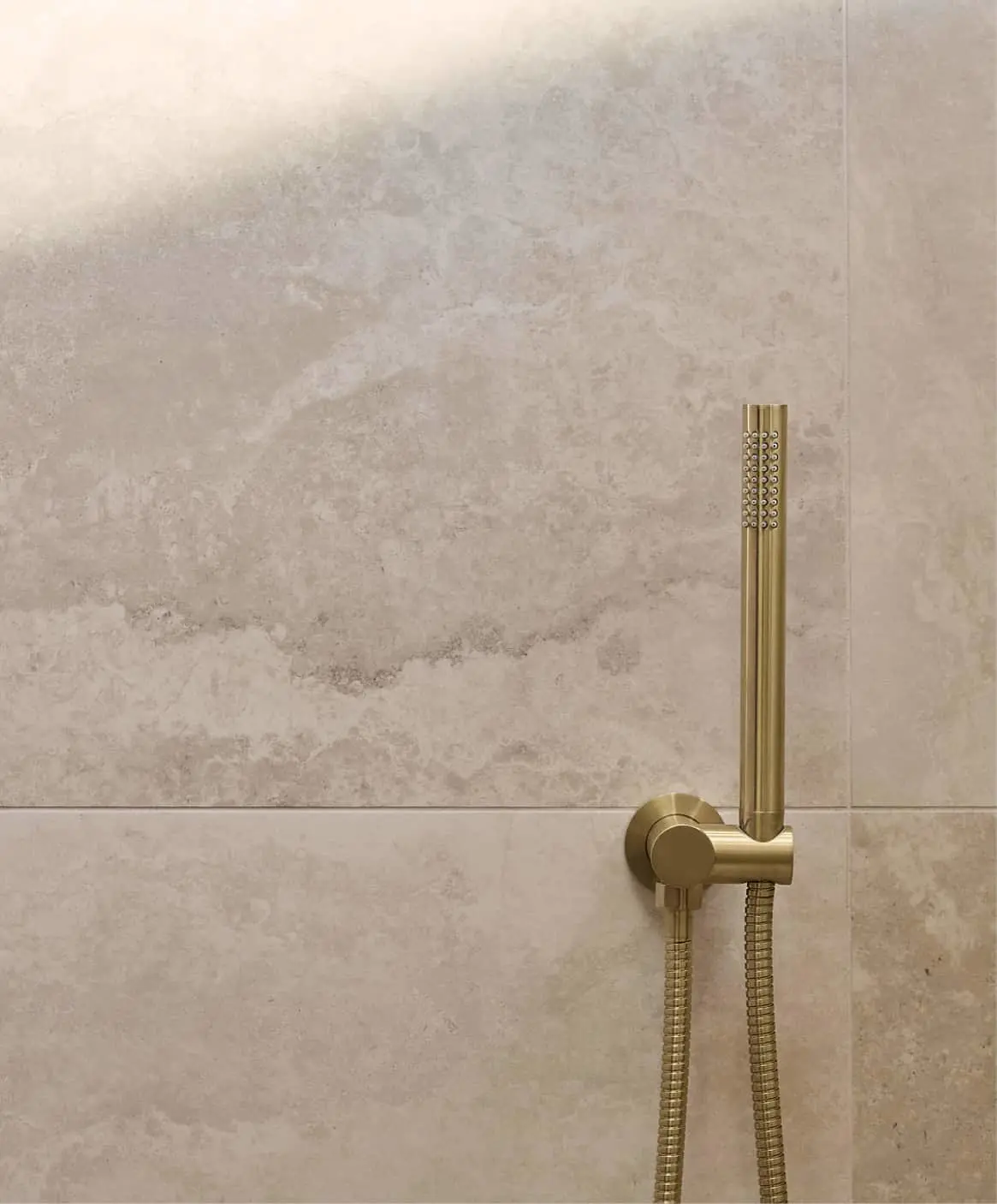 Modern shower with beige stone tiles, handheld gold sprayer, natural light from window.