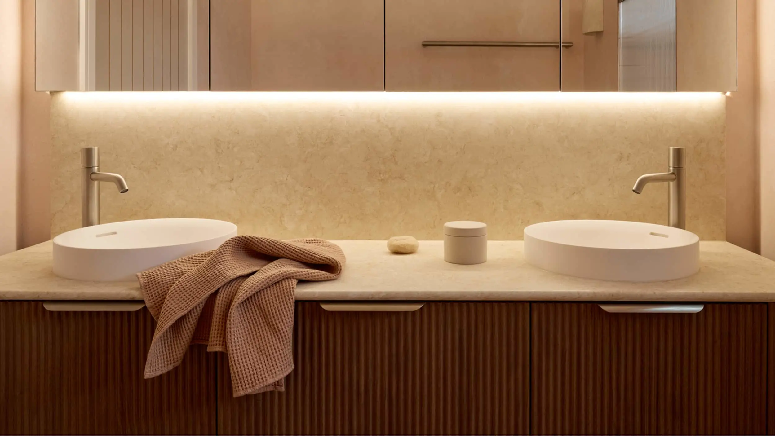 Modern bathroom vanity with dual white vessel sinks, beige countertop, wooden cabinetry, and ambient lighting.